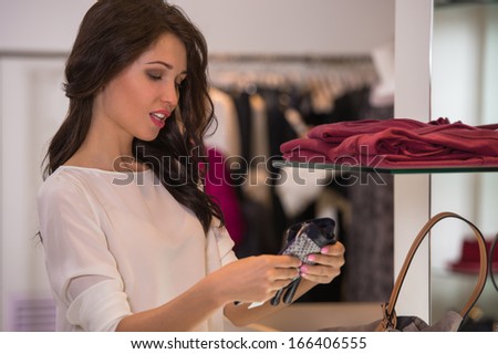 Young pretty woman at luxury boutique choosing accessories while standing near glass shelf and mirror