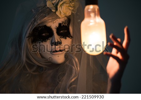 Halloween witch. Beautiful woman wearing santa muerte mask casting spell near light bulb