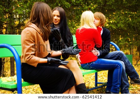 Group Of Four People Sitting On Bench In Autumn Park. Young Couple Kissing. Two Adult Women Laugh At Them.