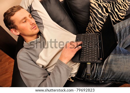 Happy young man sitting on sofa at home, working on laptop computer, smiling
