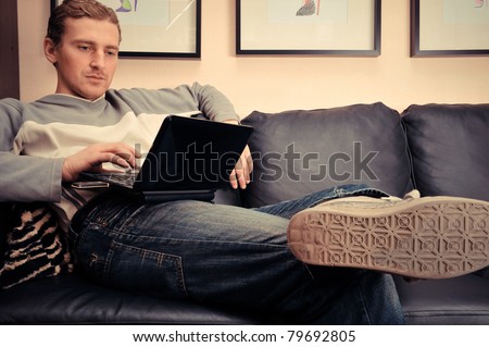 Happy young man sitting on sofa at home, working on laptop computer, smiling
