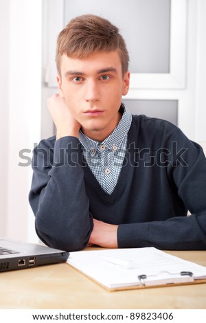 Young student man working in bright room, sitting at desk, using laptop and papers