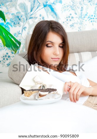 Delighted woman reading a greeting card sitting on the sofa at home and eating tasty cakes