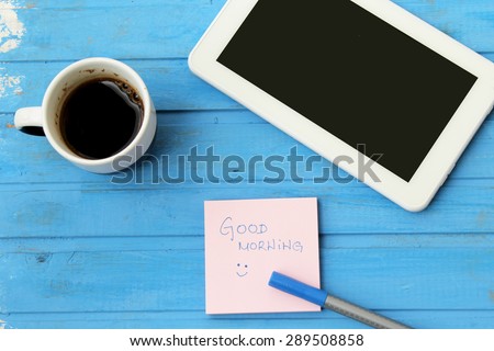 Touch screen tablet computer, cup of coffee, sticky note with good morning message and blue pen - on blue background