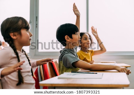 Elementary age Asian student boy raised hands up in Q and A class. Diverse group of pre-school pupils in elementary age in education building school. Volunteering and participating classroom concept.
