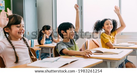 Elementary age Asian student boy raised hands up in class. Diverse group of pre-school pupils in elementary age in education building school. Volunteering and participating classroom concept.