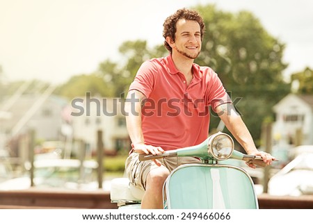Young, bearded man in red shirt and cargo shorts riding vintage restored scooter through marina.