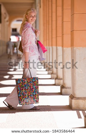 Woman on a holiday carrying shopping bags with her and walking down a sunny corridor while looking at the camera and smiling.