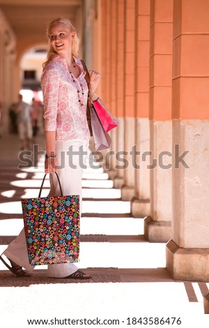 Vertical shot of a joyous woman on a holiday walking down a sunny corridor carrying shopping bags with her while looking away.