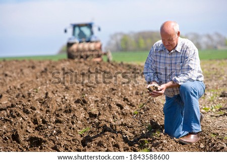 Smiling old-aged farmer kneeling and holding soil in his cupped hands with a tractor and a plough in the background