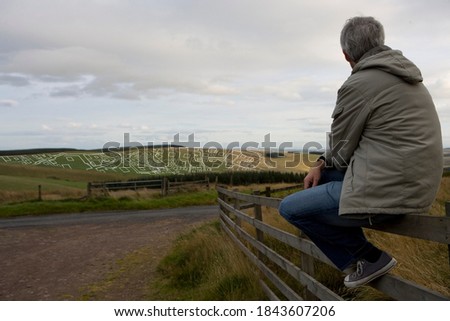 Horizontal portrait of a senior man seated on a wooden fence overlooking housing development layout in distance with copy space.