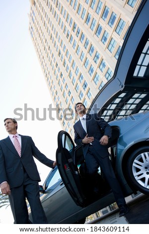Tilted low angle shot of a businessman exiting a car with the door held open by the chauffeur in front of a skyscraper.