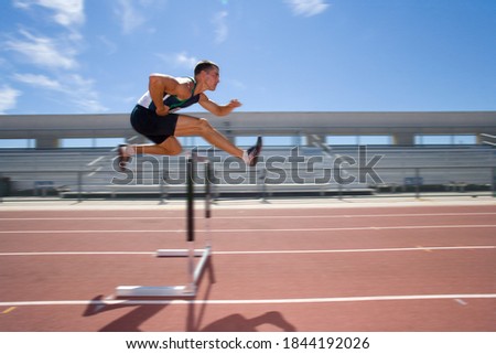 Blurred motion shot of male athlete jumping over a hurdle in the race on running track
