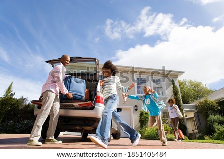 Horizontal low angle shot of two playful children running by the father packing a suitcase into a car with mother striding in the background.