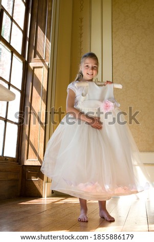 Vertical low angle shot of a girl holding up a flower girl dress standing by the window smiles at the camera.