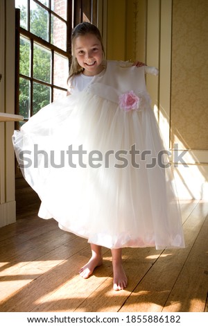 A girl holds up a flower girl dress standing by the window smiles at the camera.
