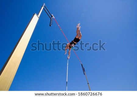 Wide shot of a pole vault athlete going over bar with a clear blue sky and lens flare in the background.