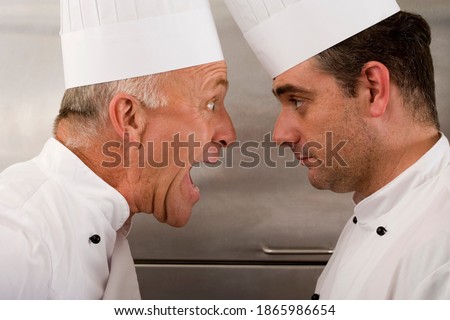 Close-up portrait of an angry senior chef yelling at the junior chef in a commercial kitchen.