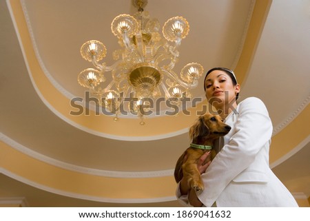 Low angle view of a young glamorous woman carrying a dog and standing below the chandelier.