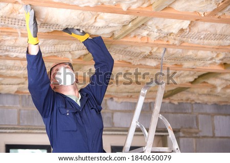 A close up shot of a man wearing protective mask standing on a ladder and installing ceiling insulation.