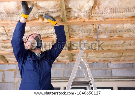 A man in coveralls wearing protective mask standing on a ladder and installing ceiling insulation in an attic.