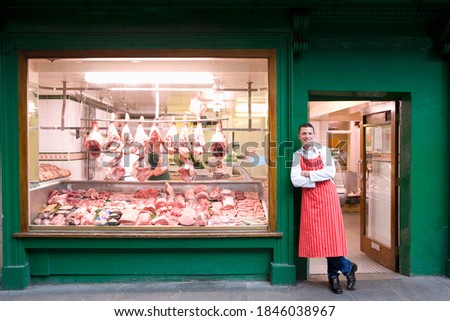 A wide shot of a young smiling butcher in red apron leaning against butcher shop doorway next to the display window. Stockfoto © 