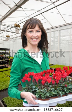 A portrait shot of a woman smiling at camera while holding a tray of flowers in a greenhouse.