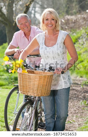 Vertical shot of a senior couple with their bicycles on a sunny day smile at the camera with the woman in front carrying spring flowers in her basket.