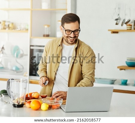 Portrait of a young man with a laptop preparing smoothie juice in blender and having fun in the kitchen at home Foto stock © 