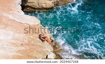 Beautiful girl sitting on a high rock and looking out to sea. top view. Girl in Hat on the edge of cliff. blue sea and high cliffs. Seascape. Girl at sunset. Sea tour. Blue sea, rocks