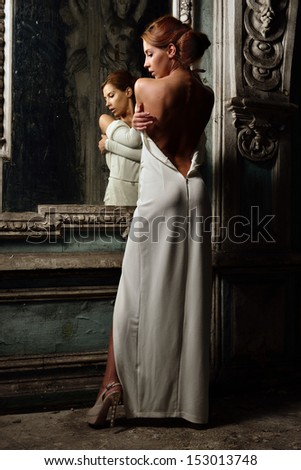 Portrait of the beautiful woman in white dress with naked back. She is standing at the mirror. Studio with interior of old palace. Not necessary property release.