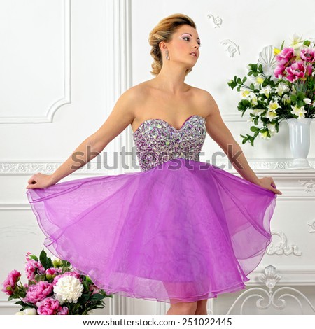 Beautiful woman in violet gown dress. White Luxury studio with flowers.