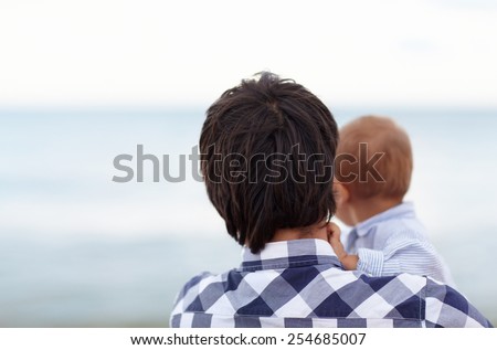 Rearview of a father holding his little baby boy, looking out to the horizon on the ocean