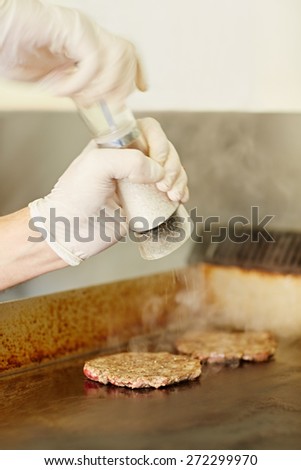 Cropped chef\'s hands wearing gloves busy grinding salt onto hamburger patties to season them while they are frying