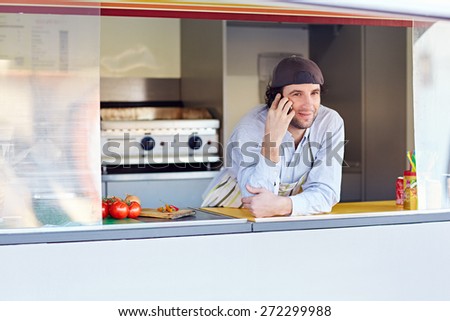Male entrepeneur leaning on the counter and talking on the phone in his food stall where he makes and sells takeaway food
