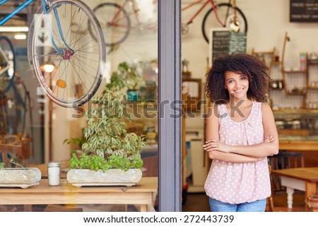 Young mixed race woman smiling while standing in the door of her cafe with her arms folded proud to be the owner of a small business