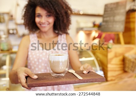 Focus on the foreground of a latte ay being carried on a wooden tray by a young mixed race waitress