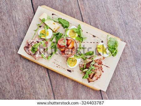 Overhead shot of a wooden platter with a fresh and healthy salad of tomato, chees, rocket and different kinds of ham, presented on a wooden table