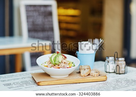 Fresh chicken and avocado salad in a presented in a bowl on a wooden board alongside some freshly sliced bread laid out on a table outside a gourmet delicatessen eatery