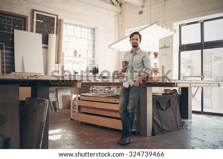 Full length portrait of a young male designer standing comfortably in his naturally lit workshop holding his morning cup of coffee and smiling at the camera