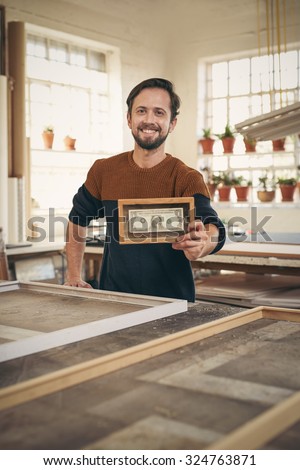 Young craftsman holding up a bank note that has been framed while standing proudly in his workshop