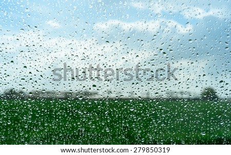 Fresh green field with the clouds in the sky behind the raindrops on the window
