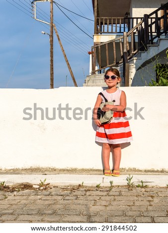 Little girl in sunglasses with a slight smile on her face dressed in a striped dress standing with a bag over her shoulder looking far away