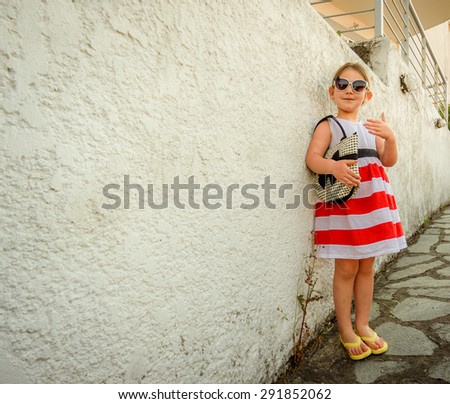 Little girl in sunglasses with a slight smile on her face dressed in a bright striped dress waving her hand