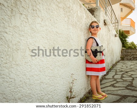 Little girl in sunglasses with a slight smile on her face and her chin raised dressed in a bright striped dress standing on the stone pathway