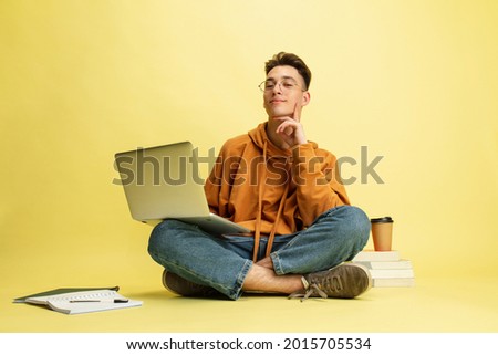 Studying, doing homework. One young smiling caucasian man, student in glasses sits on floor with laptop isolated on yellow studio background. Education, studying and student life concept. Сток-фото © 