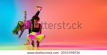 Emotive dance style. Portrait of two young beautiful hip-hop grils dancing on colorful gradient blue orange background in neon. Youth culture, movement, active lifestyle, action, street dance, ad