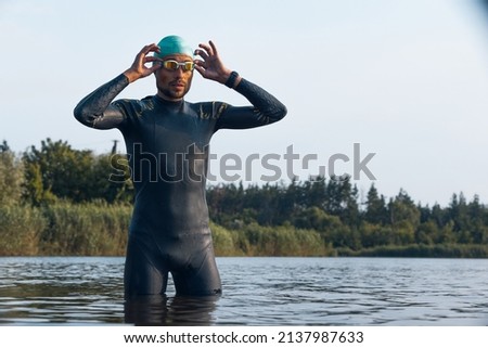 Professional triathlete before swimming in river's open water. Man wearing swim equipment practicing triathlon on the beach in summer's day. Concept of healthy lifestyle, sport, Imagine de stoc © 