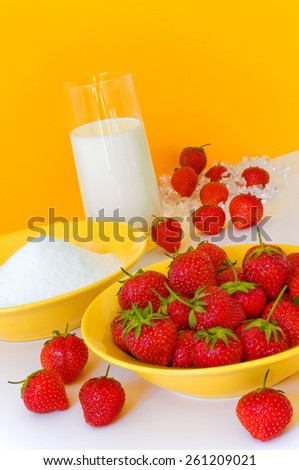 Fresh strawberries, sugar and a glass of milk arranged on white ground against yellow background, ingredients for delicious strawberry milkshake