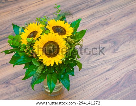Flower bouquet with three sunflowers and green leaves in glass vase on parquet floor